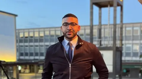 Reform UK Mason Humberstone with dark hair and glasses wearing a shirt and tie and a bomber jacket, standing in a town square and leaning against railings. 