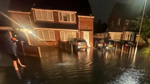 BBC Man standing in flooded road at night with houses and parked cars in background. Inches of water cover the entire road and driveways.
