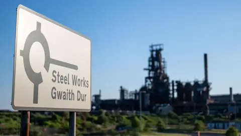 Getty Images A bilingual sign in the foreground reads “Steel Works / Gwaith Dur” beside an industrial symbol. Behind it stands a large steelworks facility with tall chimneys and complex machinery, set against a clear blue sky. 