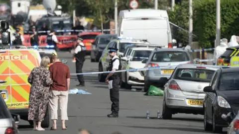 Getty Images Members of the public look on from in front of blue and white police tape as police officers stand in a road filled with police vans and cars, with debris strewn across it.