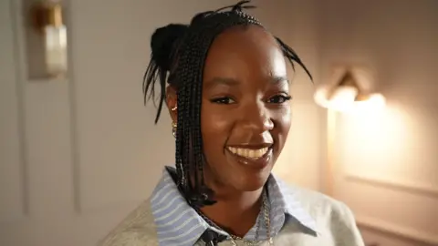 BBC Corinna Brown is seen smiling widely in a portrait photograph. She is facing the camera and is wearing a grey jumper over a blue striped shirt