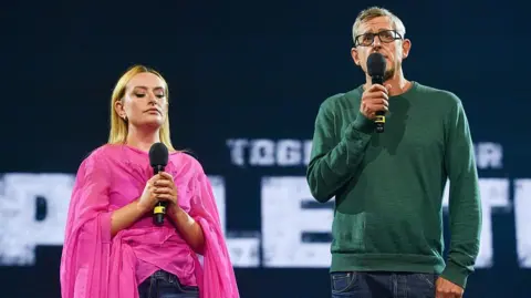 Getty Images Amelia Dimoldenberg and Louis Theroux stand in front of a Together for Palestine banner. Both hold microphones, Louis is speaking into his while Amelia holds hers at chest level, listening intently.
