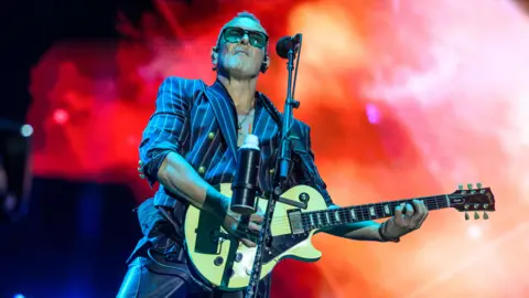 Getty Images Vivian Campbell of Def Leppard performs onstage. He's holding a guitar and wearing a navy stripped blazer with gold buttons and sunglasses. Lights behind him are red.