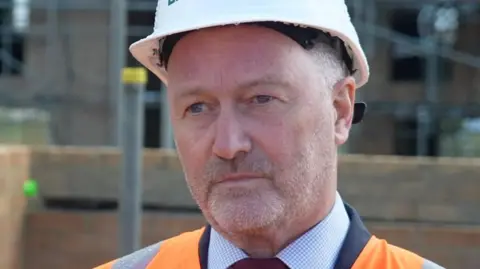 BBC Housing Secretary Steve Reed wearing a white hard hat and orange hi-vis jacket.