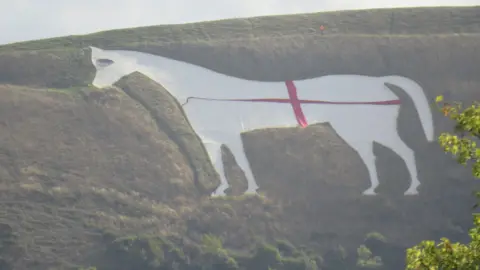 BBC A zoomed in picture of a large white horse carved on the side of a hill in Wiltshire. There is red fabric draped across it in a cross shape. 