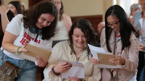 PA Photo shows three girls collecting their GCSE results. They all have long, dark hair and are smiling down at their results papers. One of the girls wears a t-shirt which says 'don't be jealous', one is a wheelchair user and one is wearing glasses.
 
 