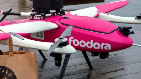 Aviant The foodora drone is pink and white with rotors on mounts attached to the wings - a third propeller is on the back. In the foreground is a brown paper bag from Bastard Burgers.