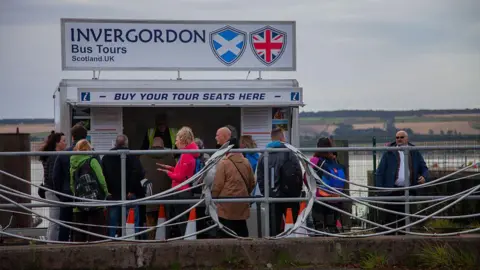 Getty Images A queue of people outside a kiosk advertising bus tours in Scotland. They are all wearing jackets on quite a grey day. The kiosk is next to the water. 