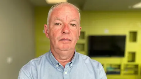 Gordon Mackenzie looking directly at the camera. He is wearing a blue shirt and has short grey hair. There is a yellow wall in the background with a wall-mounted TV and some cut-out sections for storage.