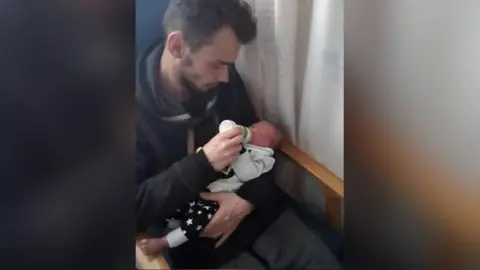 North Wales Police/PA Wire A man with brown hair holding a baby in his left arm. With his right hand, he is feeding the child with a bottle of milk. They are sitting in a chair with a curtain visible on the left of the pair of them.