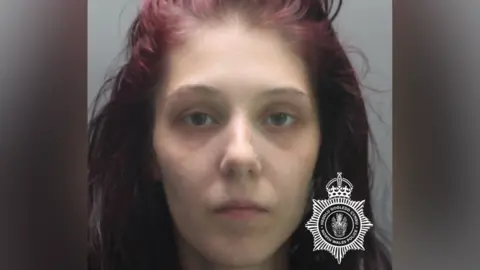 North Wales Police A mugshot of a woman with died red-purple hair. She is pale with a nose stud and is looking directly at the camera