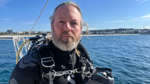 A man with grey hair and a grey beard looks at the camera. He is wearing a wetsuit and diving equipment. The ocean and a beach can be seen in the distance.