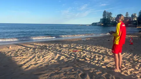 A blonde man dressed in a bright red and yellow lifeguard uniform stands on the beach and flies a drone.