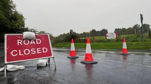 A red sign that says road closed and three bright orange traffic cones sit on a road