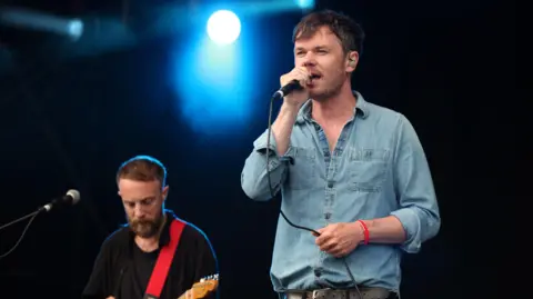 A man wearing a blue denim shirt and with short dark hair singing into a microphone. Another man is in the background playing guitar.