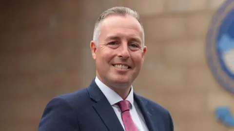 PA Media Paul Givan is standing in front of a light brick wall and is smiling at the camera. He is a middle-aged man, with short, greying hair. He is wearing a navy blazer jacket, a white collared shirt and a red paisley tie.
