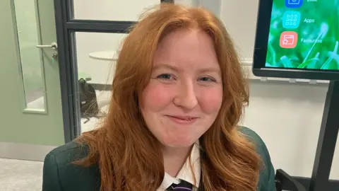 A teenage girl with long ginger hair, a green blazer and tie smiles at the camera.