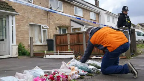 PA Media A man in an orange coat and jeans placing flowers on paving slabs outside a light brick house with police tape outsider and a police officer in full black uniform stood near by