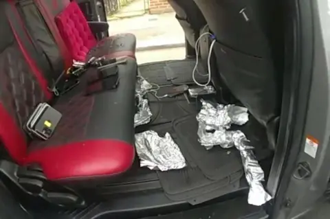 Police bodycam footage shows multiple mobile phones are seen on the back seats of a grey car, with wraps of foil strewn across the floor of the car. The seats are red and black.
