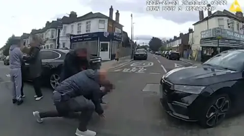 Police bodycam footage shows an plain-clothes office restraining a man on the ground in the middle of the road after unmarked cars intercepted a car suspected to have been transporting stolen phones. The unmarked officer is wearing a navy blue coat and grey trousers, next to a black unmarked police car.