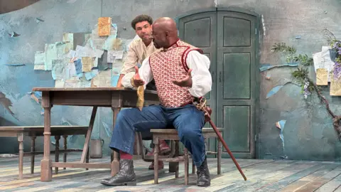 Two men performing on stage during a play. One has an enhanced and disfigured nose and is sitting at a bench, next to a table, with his arms outstretched as he speaks to another man over his shoulder.