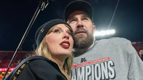 Getty Images Taylor Swift stands beside her fiance Travis Kelce inside an American football stadium. She is wearing a black hoodie with a black hat on. He is wearing a grey jumper with the word 'champions' on it and a black cap.