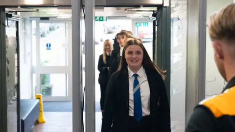Pupils walk through a metal detector a member of the school's staff stands in the foreground.