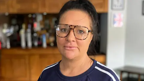 Caroline is wearing big glasses and a blue sports top, she is standing in her kitchen.