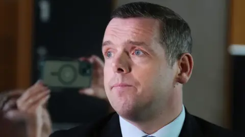 PA Media Douglas Ross, who has short dark hair, with his face lit up by a light from a camera out of shot. He is wearing a dark suit and white shirt. 