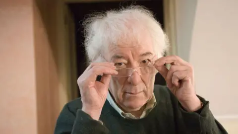 Getty Images A man with white fluffy hair holds his glasses on the end of his nose. He looks at the camera with a neutral expression. 