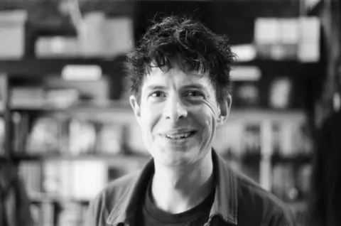 Stephen Connolly A black and white image of a man smiling at the camera. He has dark hair and is wearing an open shirt. 