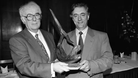 Independent News and Media/Getty Images A black and white photo of a white haired man with black rimmed glasses, wearing a suit, being presented with a sculpture by a man with dark hair in a suit. 