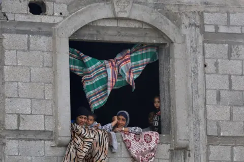 Reuters Women and children peer out of the window of a stone building. There are rugs draped over the window and hanging from the ceiling.