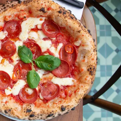 Catriona Byers The pizza is topped with cheese, tomato sauce, pepperoni and basil. It is on a plate on a table next to a napkin with a knife and fork.