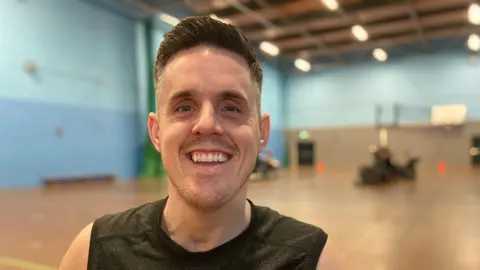 A man sat in a sports hall is smiling at the camera, he is wearing a grey vest. 