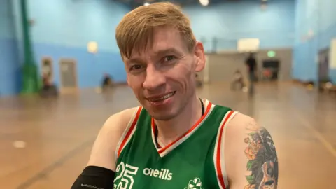 A man with red hair looks at the camera. The picture is taken in a sports hall, and he is wearing a green vest, and has a tattoo of flowers and a guitar on his left shoulder. 
