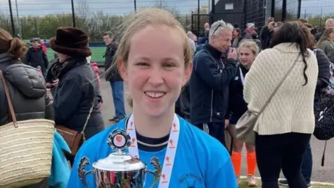 Suffolk Police Georgia Scarff in a blue sports kit, holding a silver trophy. She has a medal around her neck. Behind her are a number of adults meeting their children after a sports game. 