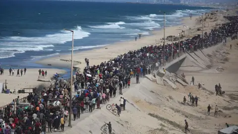 Anadolu via Getty Images Hundreds of Palestinians walk along a narrow coastal road to the north of the Gaza Strip. Most are walking on foot with their belongings. To their left is the beach and the sea. Some others are walking off road on a slope to the road's right.