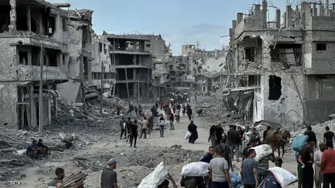 Anadolu via Getty Images People walk with their belongings in the Khan Younis area of Gaza, surrounded by homes that are extremely damaged. Rubble is strewn on the street. 