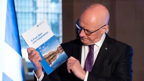 PA Media John Swinney - in the same outfit as the previous photo -  stands holding the glossy booklet  and looking at the front of it which says: "A fresh start to independence". There is a window and a Saltire in the background