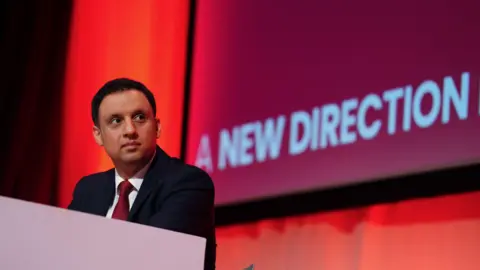 PA Media Anas Sarwar, a clean-shaven man with short black hair, a navy suit and red tie sits at a desk at a Labour conference, a red screen behind him displaying the words "a new direction".
