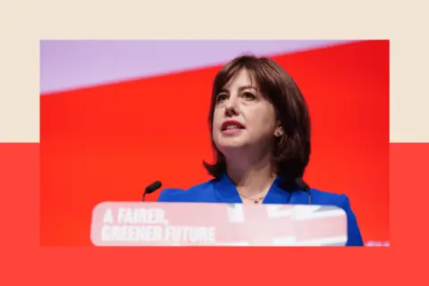 PA Wire A close up of Lucy Powell, standing at a Labour-themed lectern with the union jack flag printed on it, wearing a blue blazer and in front of a bright red background.