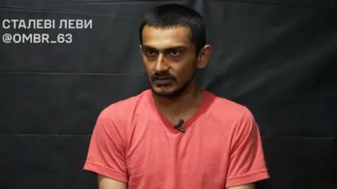 Ukrainian army/Facebook A man wearing a salmon pink shirt looks into the camera. There is a black curtain behind him. 