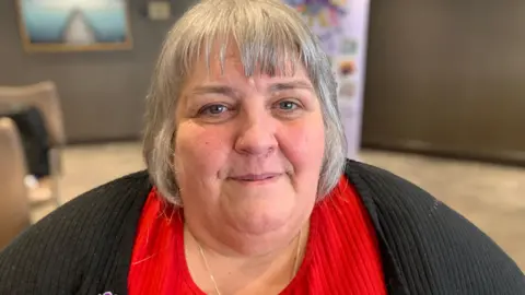 A woman with chin length grey hair is smiling at the camera. She has a black cardigan on over a red coloured top.