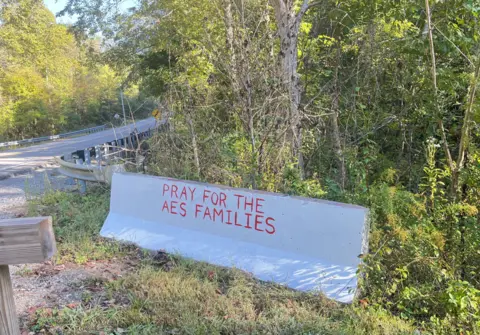 A concrete barrier on the side of a road reads "Pray for AES families".
