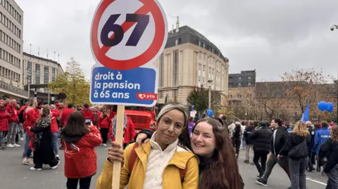 Bruno Boelpaep/BBC A young woman and her friend pose for a picture with a sign showing the number 67 with a red bar through it