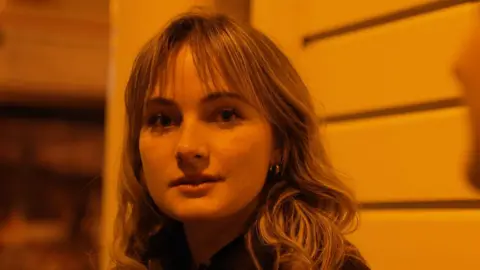 A blonde haired woman looks straight at the camera wearing earings