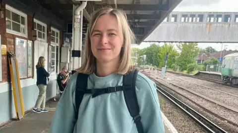 Maddie Waktare A woman with shoulder length blonde hair wearing a green jumper and a backpack standing at a train station with the railway track and a platform behind her. It is a head and shoulders shot of her.