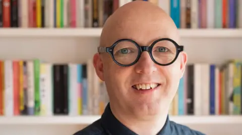 Rónán Hession A man stands in front of three shelves from a white bookcase. He wears a black shirt and green tie. He is bald and wears black clear-lensed glasses.