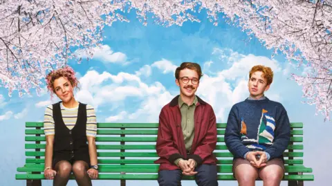 Key Art A woman with long pink curly hair sits to the far left of a green bench. She wears a black yellow and blue stripy t-shirt under a black buttoned dress. Behind her is a partially cloudy blue sky with pink blossom trees either side of the bench, hanging over it. To the right of the bench are two men. The man in the middle has short brown hair with red clear spectacles, he wears a green shirt, red jacket and navy jeans. The man on the far right wears a navy sweater with an illustration of a yacht on it. the yacht has a green white and blue sail. The man also wears a pair of brown shorts. 